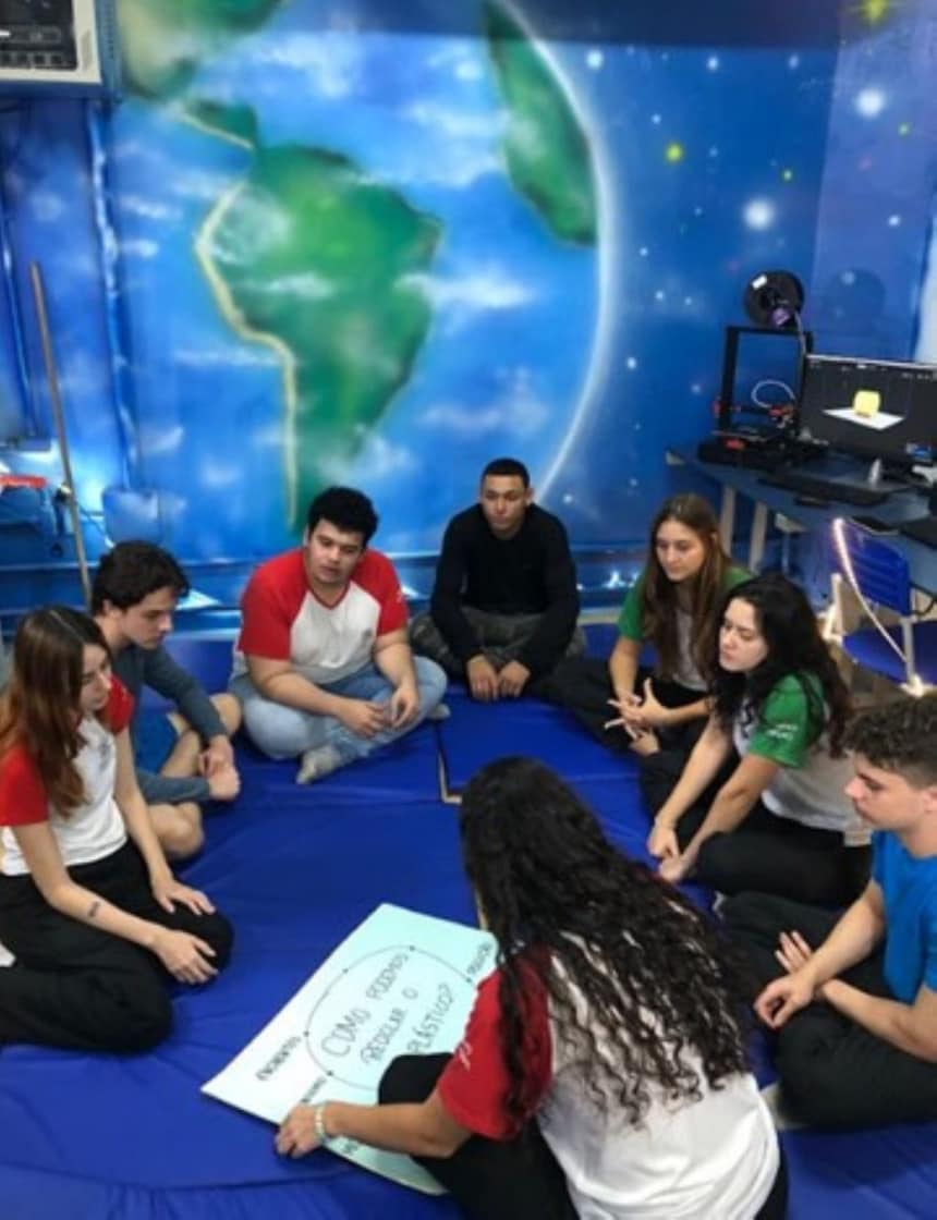 A imagem mostra os alunos sentados em um tapete azul em uma sala com paredes decoradas para se assemelhar ao espaço sideral, apresentando imagens da Terra e de estrelas. Um estudante está segurando um quadro branco com as expressões químicas "CH₄ + 2O₂ → CO₂ + 2H₂O" e "C₆H₁₂O₆ + 6O₂ → 6CO₂ + 6H₂O", que representam a combustão do metano e a respiração da glicose, respectivamente.
