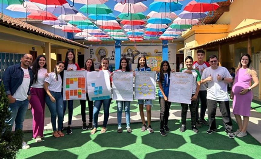 A imagem mostra os alunos e professores em pé ao ar livre sob um dossel de guarda-chuvas coloridos. Eles seguram cartazes do projeto. O cenário inclui um prédio com um portão aberto ao fundo e grama artificial no chão, marcada com a sombra dos guarda-chuvas instalados acima.